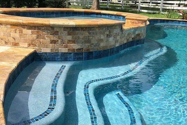A swimming pool with tiered steps featuring blue mosaic trim, adjacent to a raised stone spa.