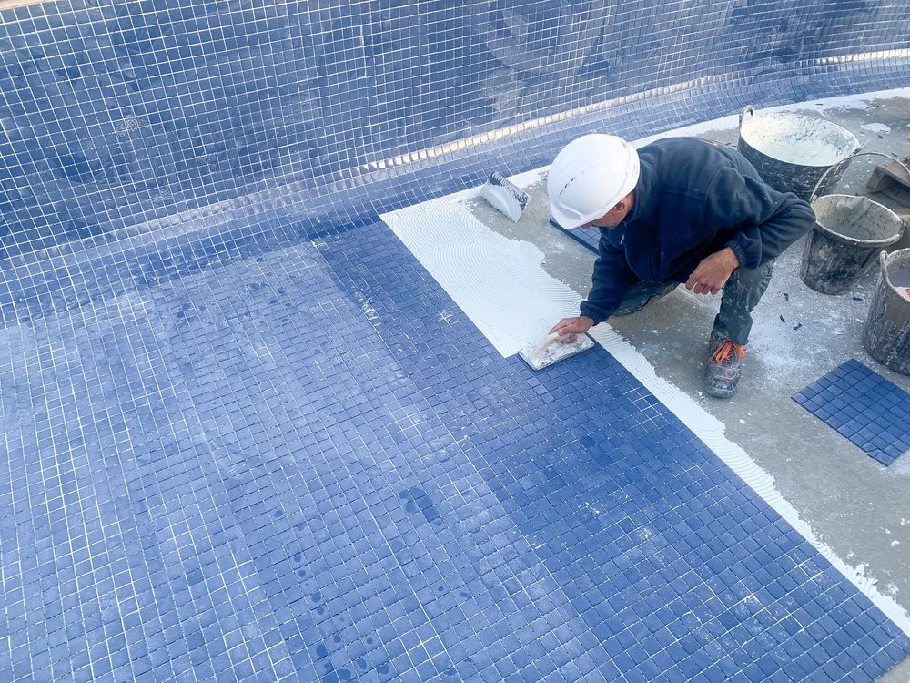 A person wearing a hard hat works on tiling the floor of a blue tiled swimming pool.