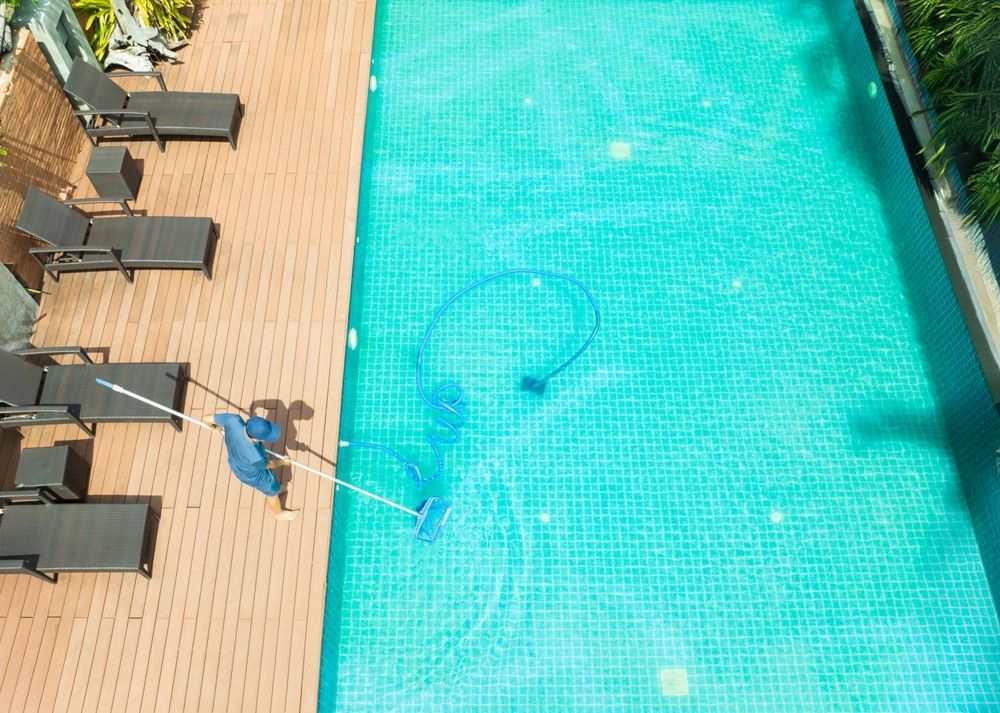 A person cleans a rectangular, turquoise-tiled swimming pool using a long-handled brush from a wooden deck with lounge chairs.