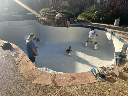 Two workers apply a white coating to the interior of an empty, sunlit swimming pool with stone coping.
