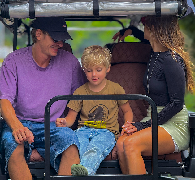 Three people sit in a golf cart, one child in the middle, outdoors under a canopy.