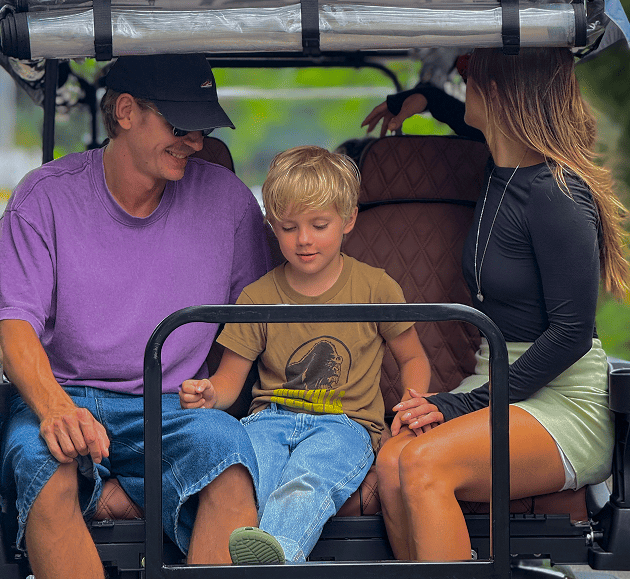 Three people seated in a golf cart, smiling and talking, with a child in the middle.