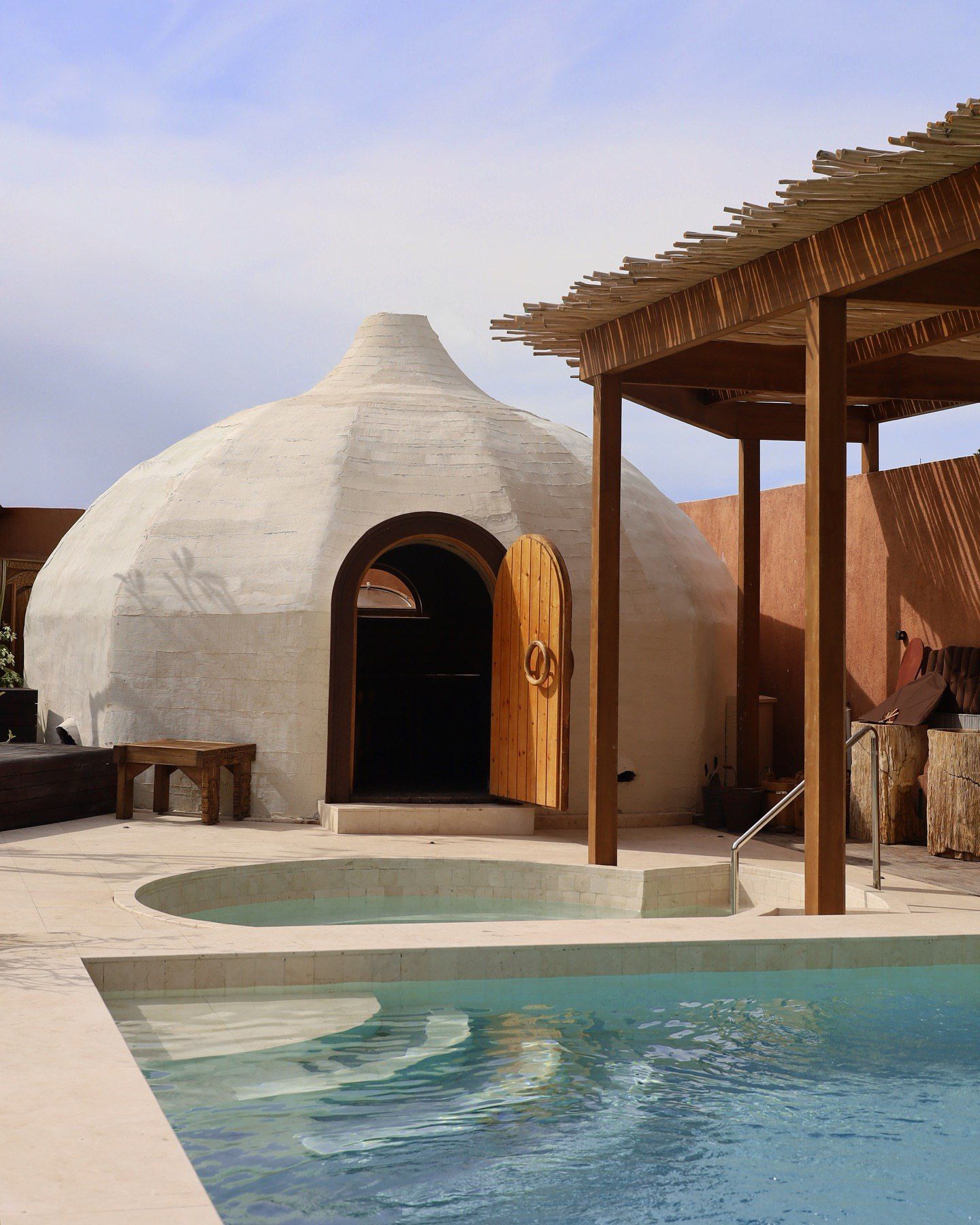 Courtyard with white dome hut, wooden pergola, and turquoise pool under a clear sky