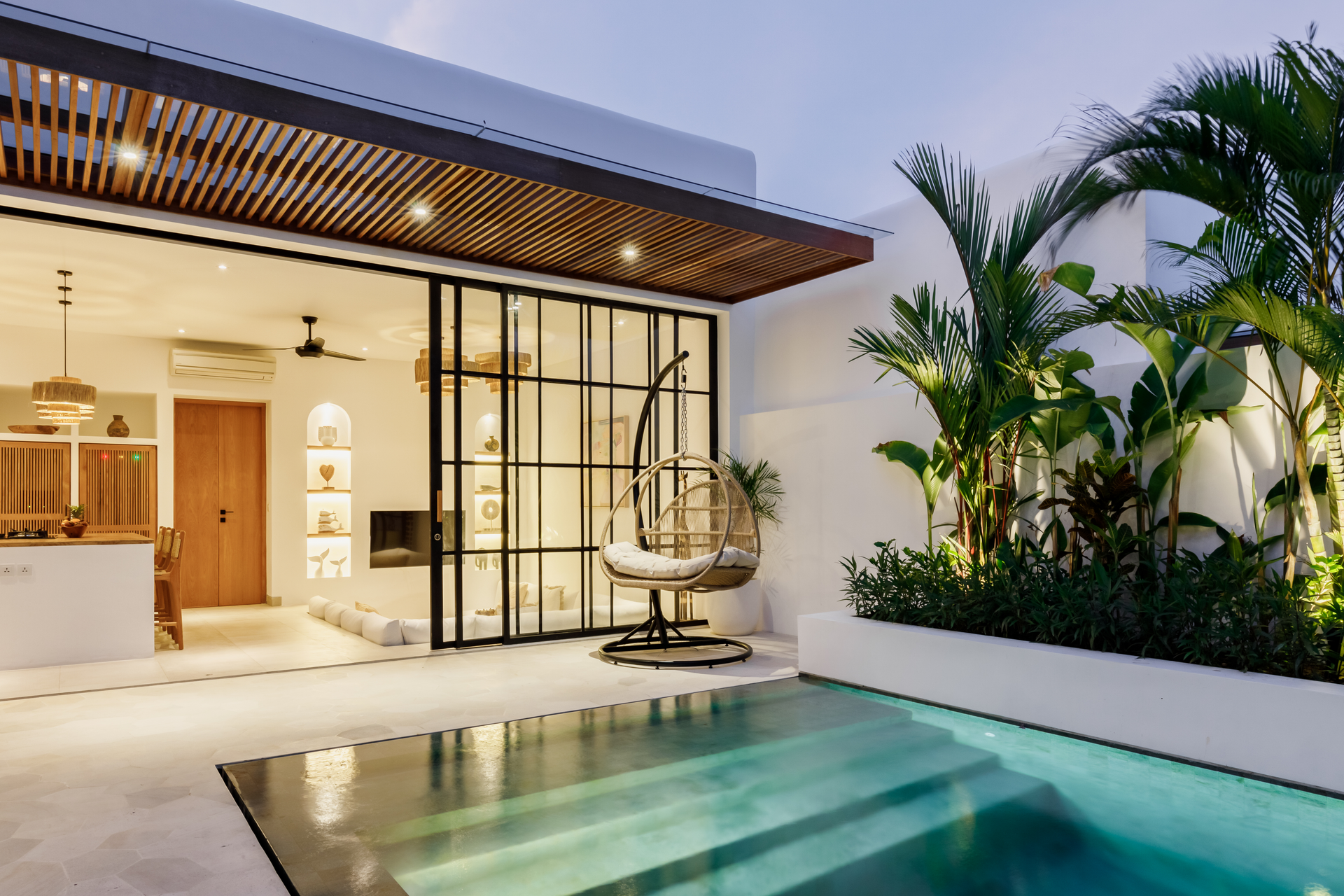 Modern tropical home courtyard with a pool, glass doors, warm lighting, and lush plants