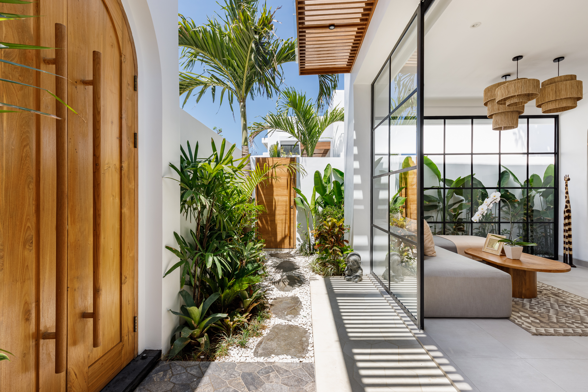 Tropical patio with wooden door, lush plants, and sunlit dining area behind glass walls