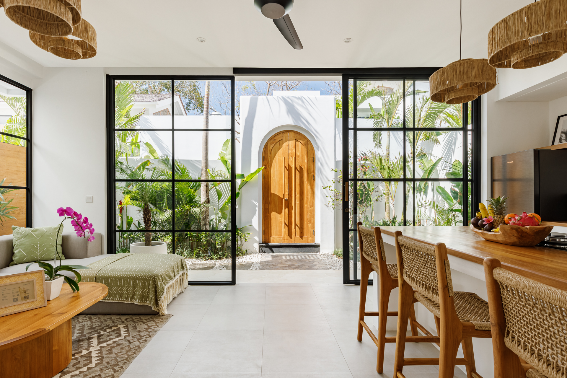 Bright dining room with wooden table, woven chairs, and large windows framing a garden view and arched front door