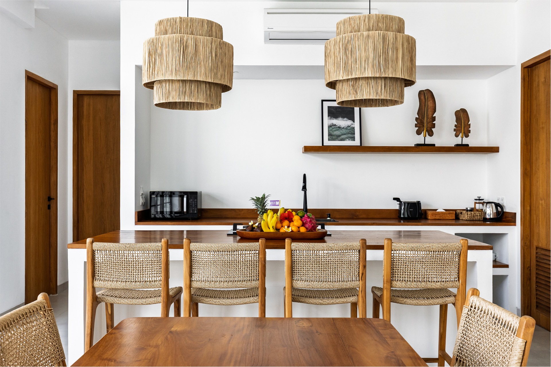 Bright dining room with wicker chairs, wooden table, hanging pendant lights, and open shelving with decor