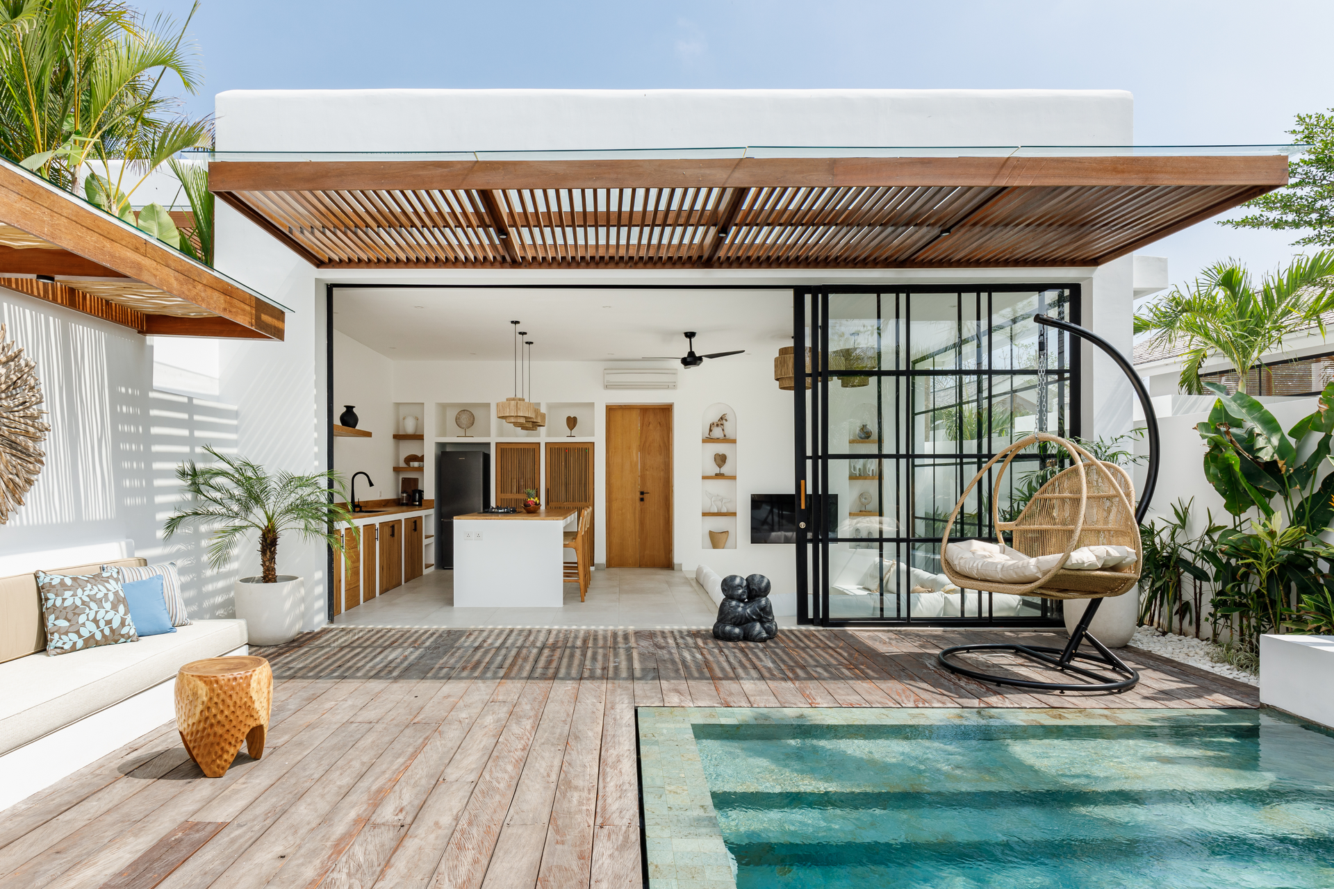 Modern patio with open kitchen, lounge chair, pool, and wood deck in a bright tropical setting