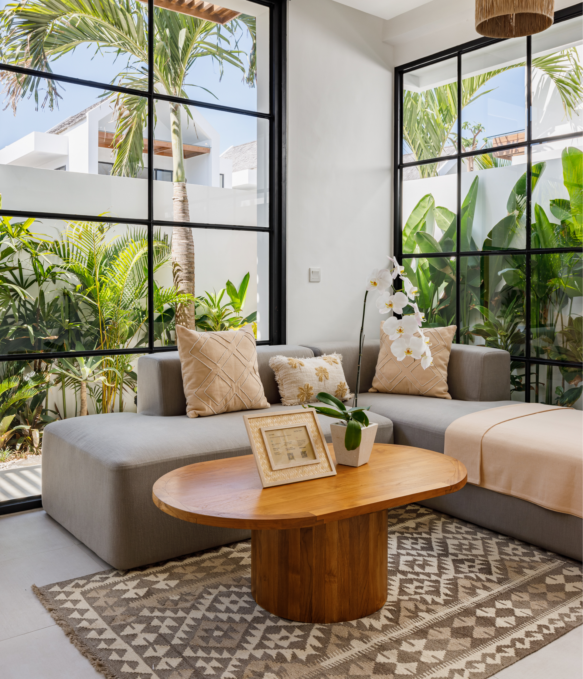 Bright living room with large windows, sectional sofa, round wood table, and lush green plants.