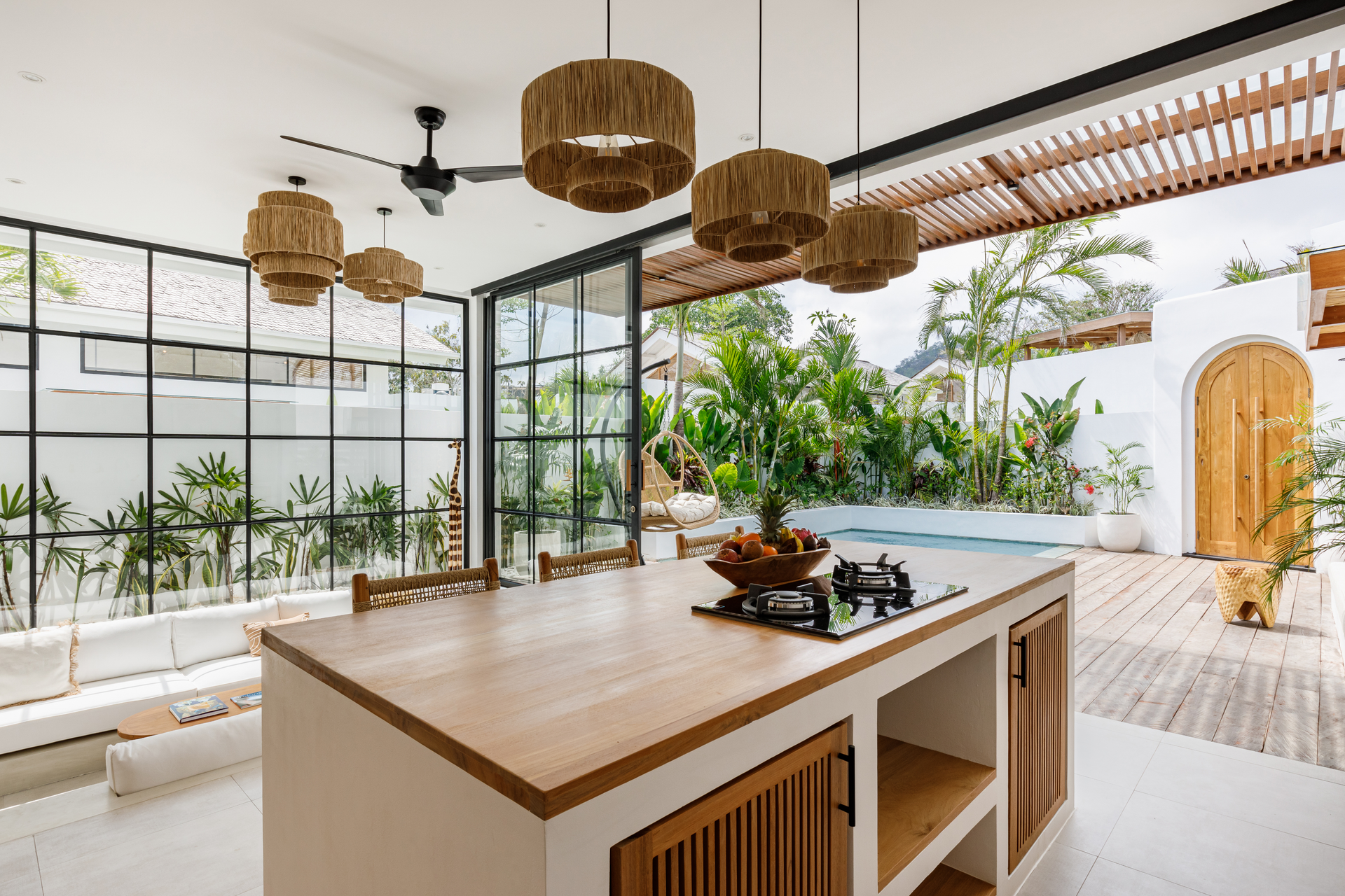 Modern open kitchen with wood island, wicker pendant lights, and large windows overlooking lush greenery.