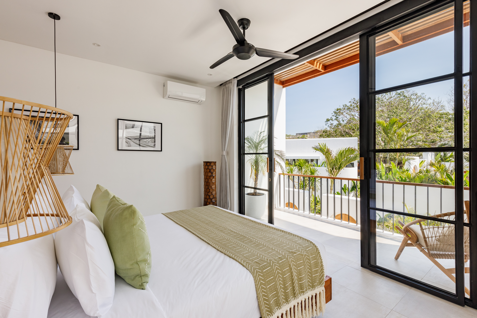 Bright bedroom with white bed, green accents, woven chair, and open doors to a balcony and tropical view.