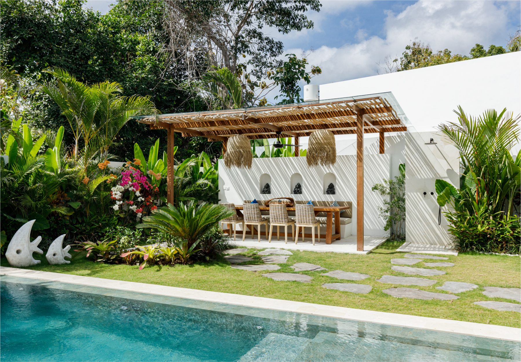 Tropical poolside patio with wooden pergola, dining table, white chairs, lush plants, and stone path
