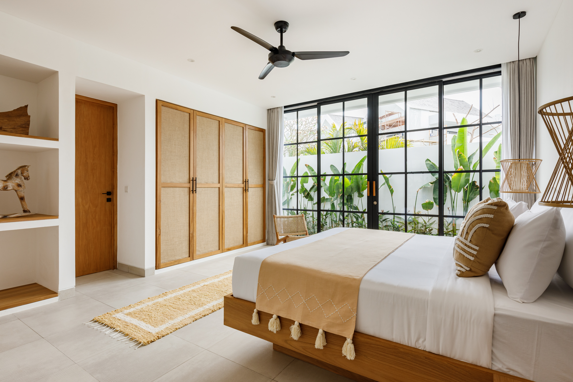 Bright modern bedroom with a wooden bed, black-framed windows, ceiling fan, and large indoor plants.