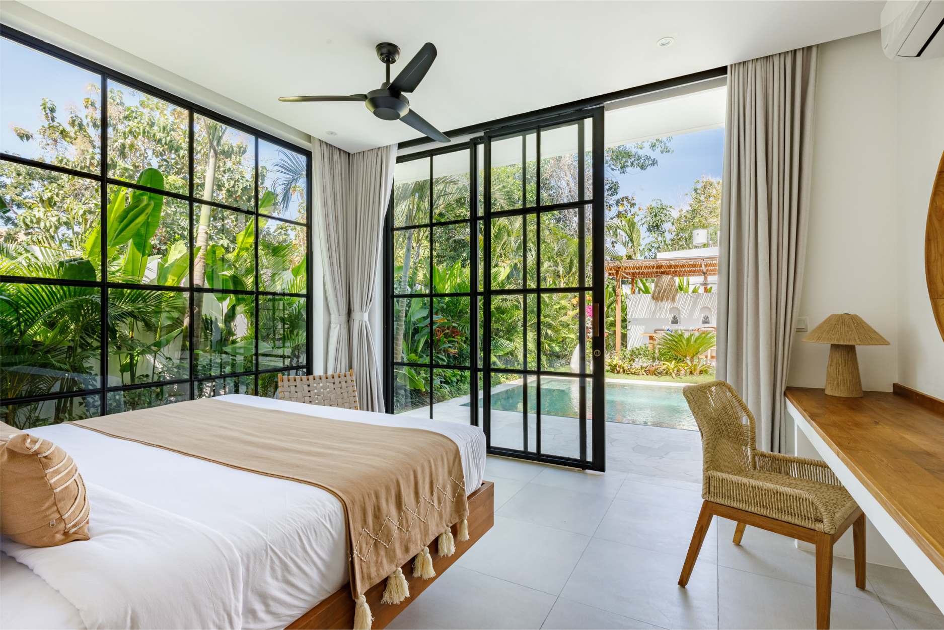 Bright bedroom with black-framed windows, white curtains, a bed, wicker chair, and garden view