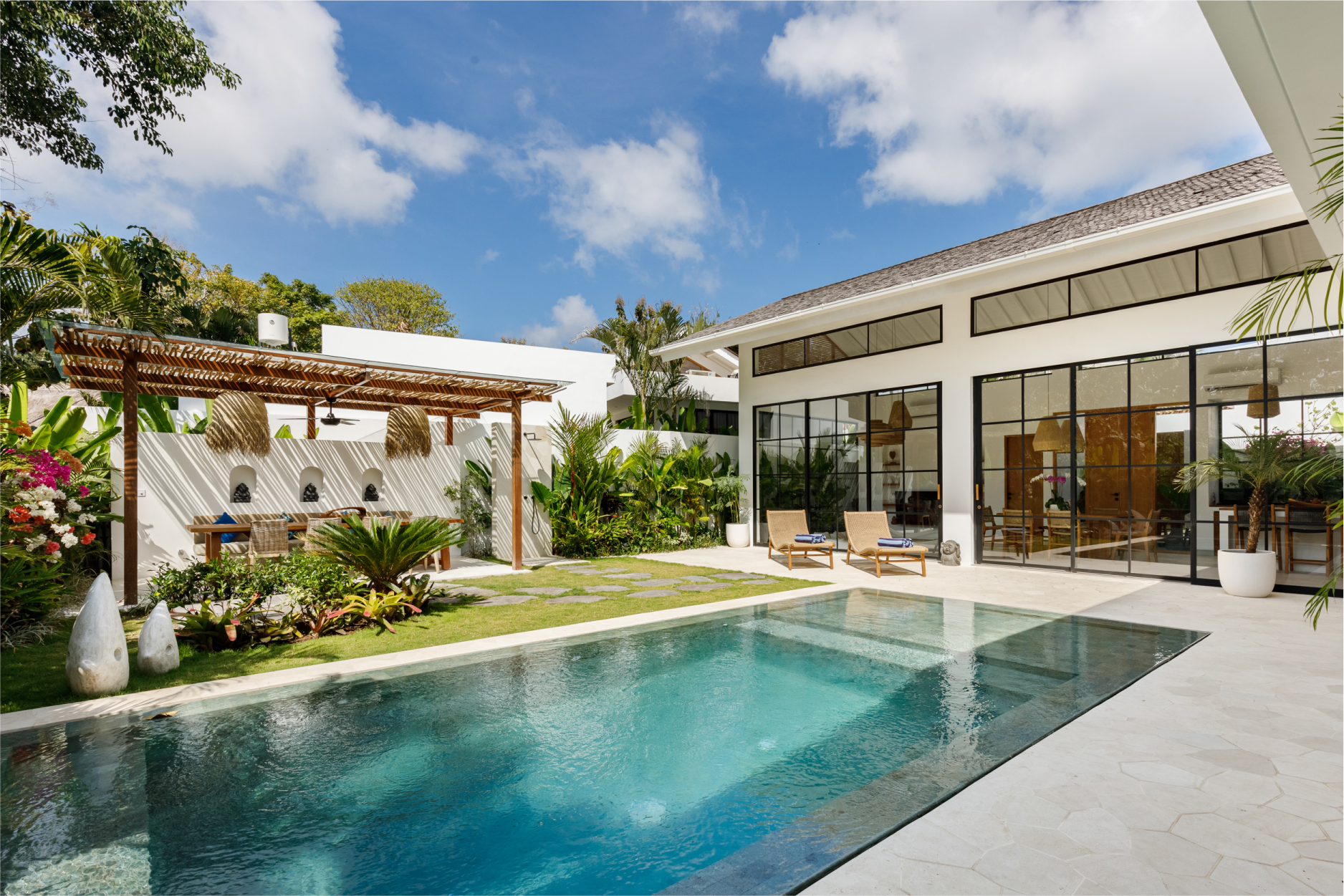 Modern villa with a turquoise pool, patio seating, and lush tropical landscaping under a blue sky