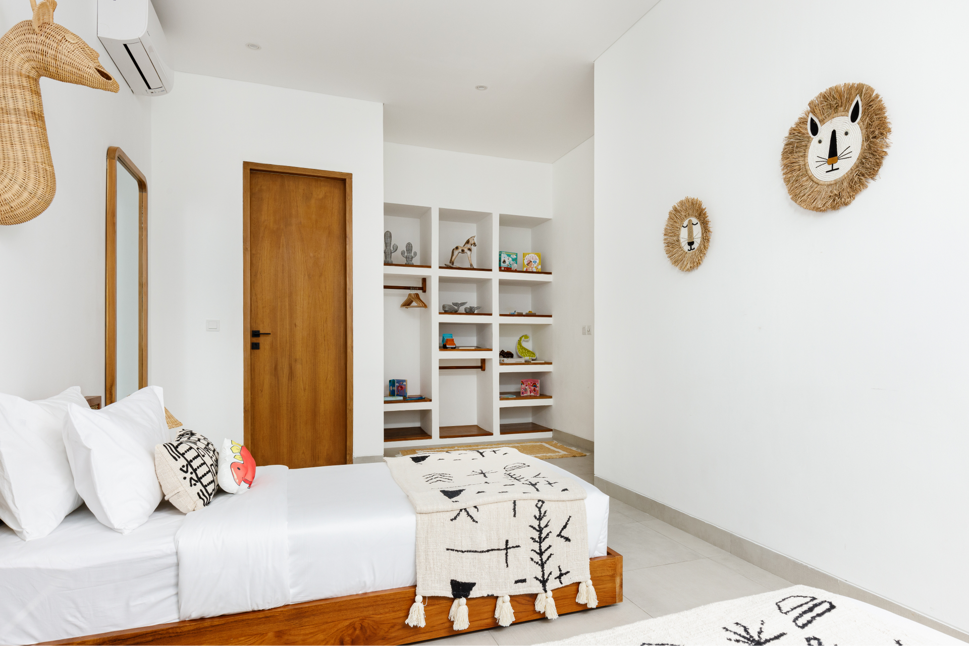 Bright minimalist bedroom with white walls, wooden bed and door, woven wall mirrors, and open shelving.