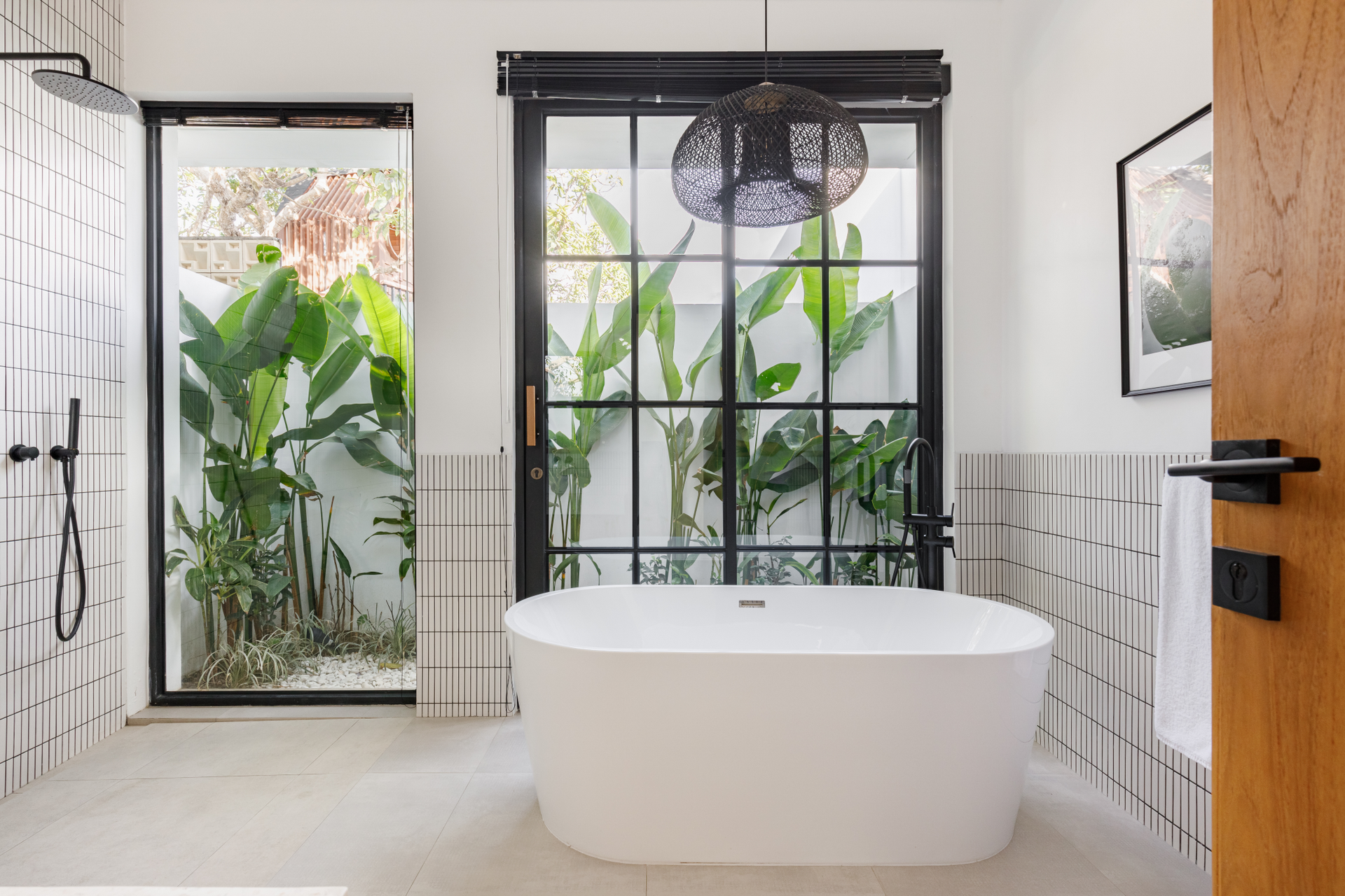 Modern bathroom with a white freestanding tub, black-framed doors, and tropical plants outside.