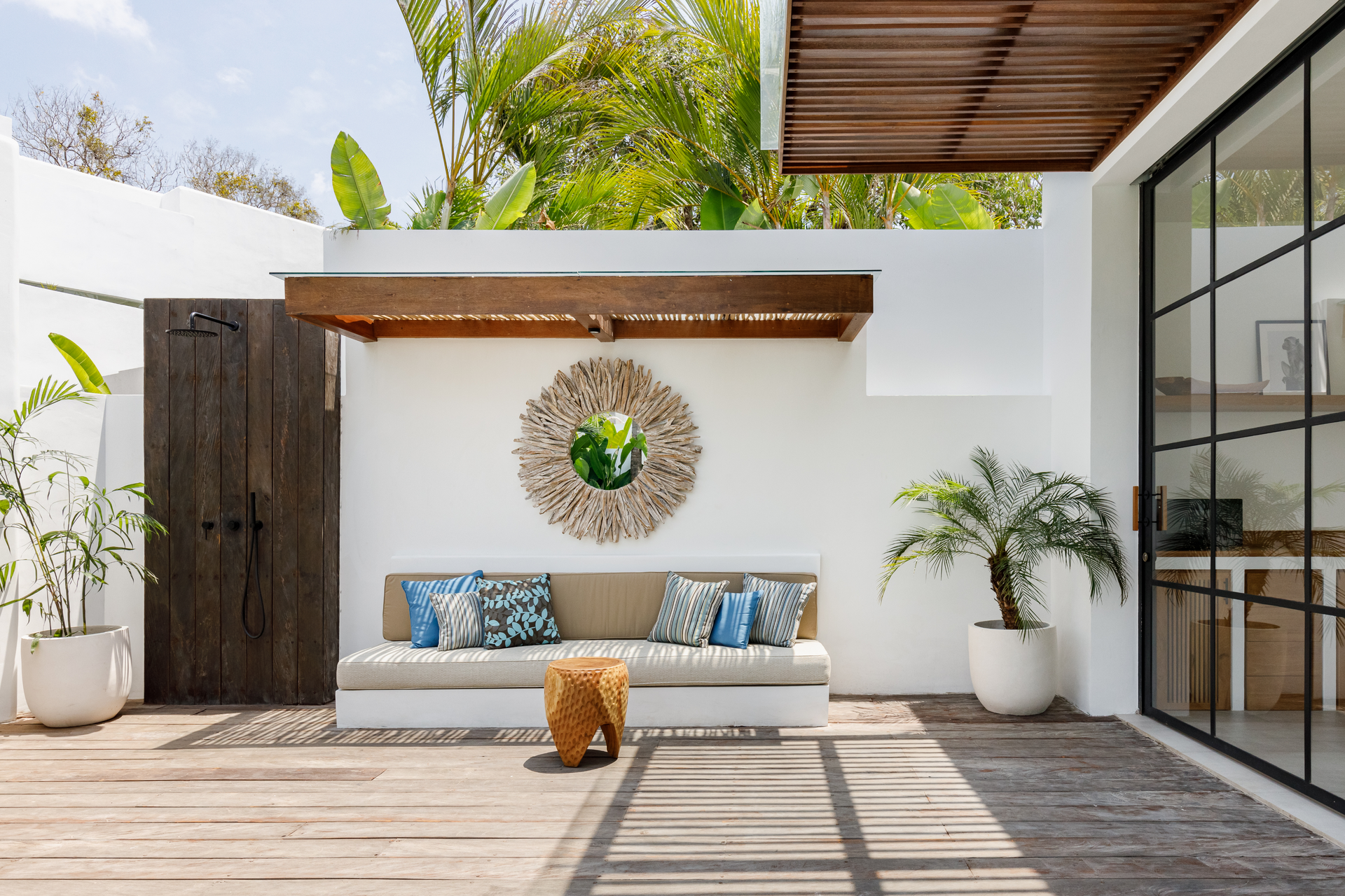 Modern patio lounge with white sofa, blue pillows, potted plants, and wooden deck under a pergola