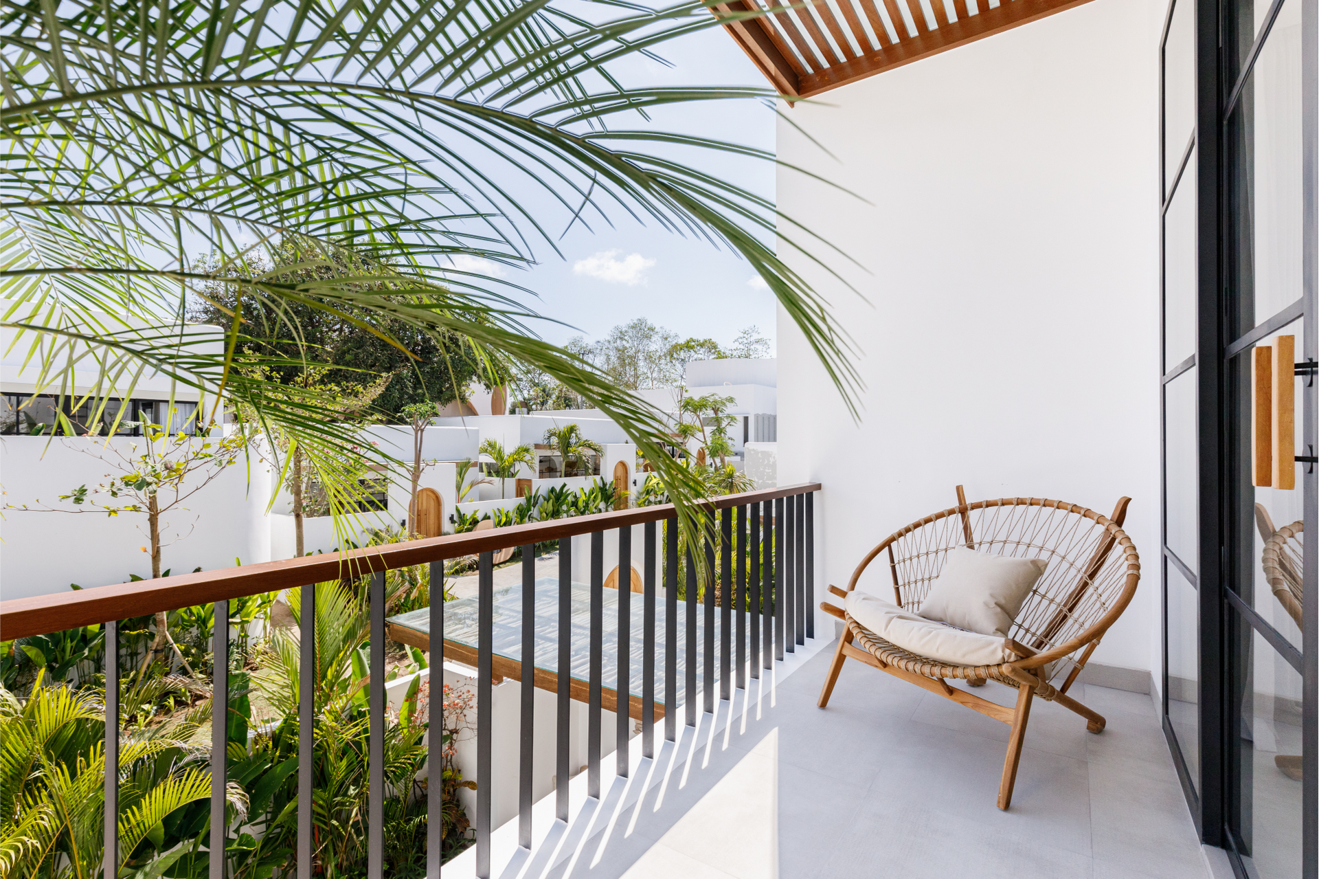 Bright balcony with a wicker chair, wooden railing, white walls, and tropical greenery beyond.