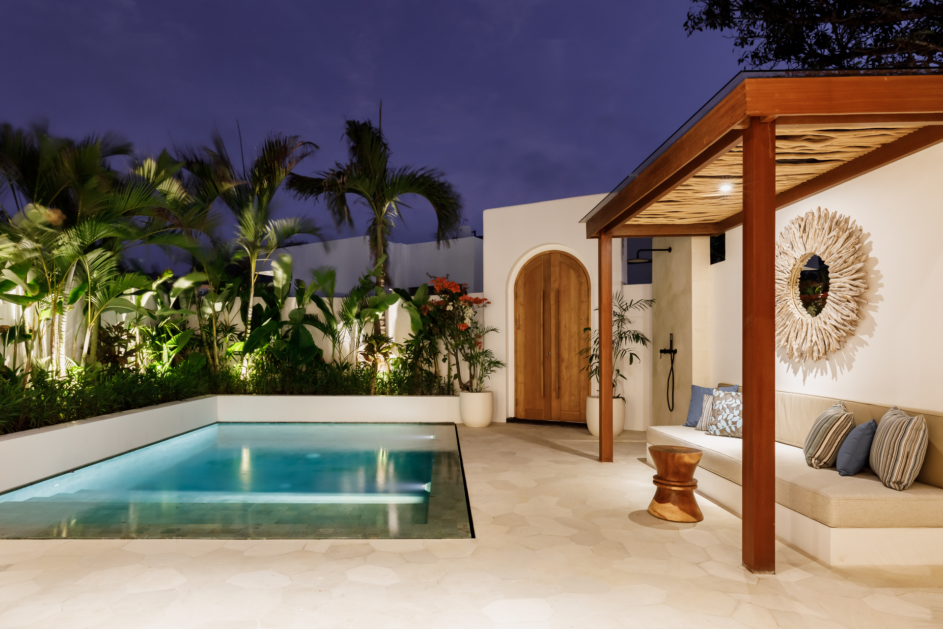 Modern patio with a lit pool, lounge seating, and tropical plants at dusk