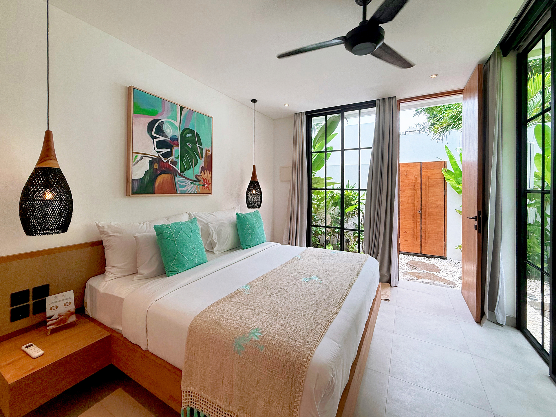 Bright bedroom with a bed, wooden nightstand, ceiling fan, and open doors to a balcony with greenery.