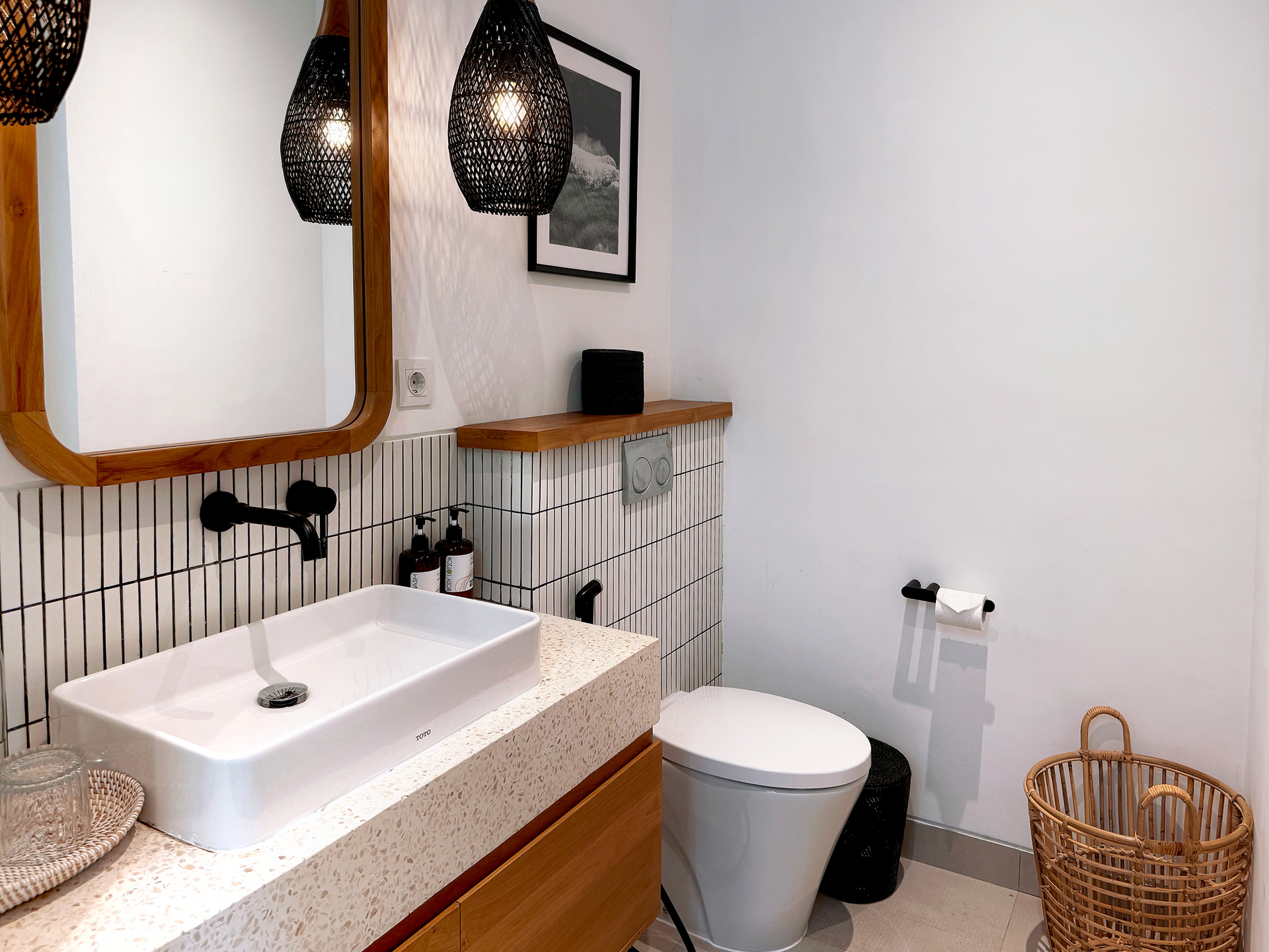 Modern bathroom with white sink, toilet, woven basket, and black pendant lights beside a wood-framed mirror