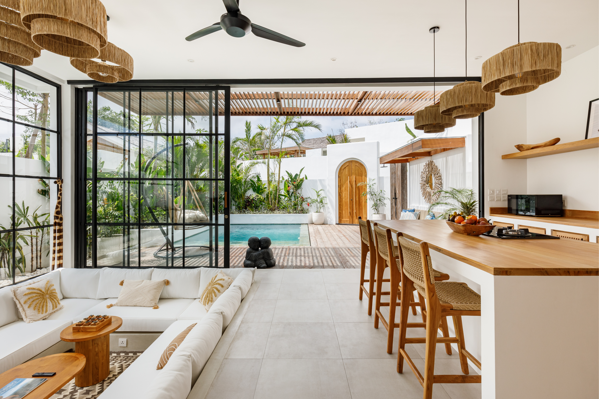 Bright open living room with white sofa, woven lights, wood bar stools, and view of a pool through glass doors