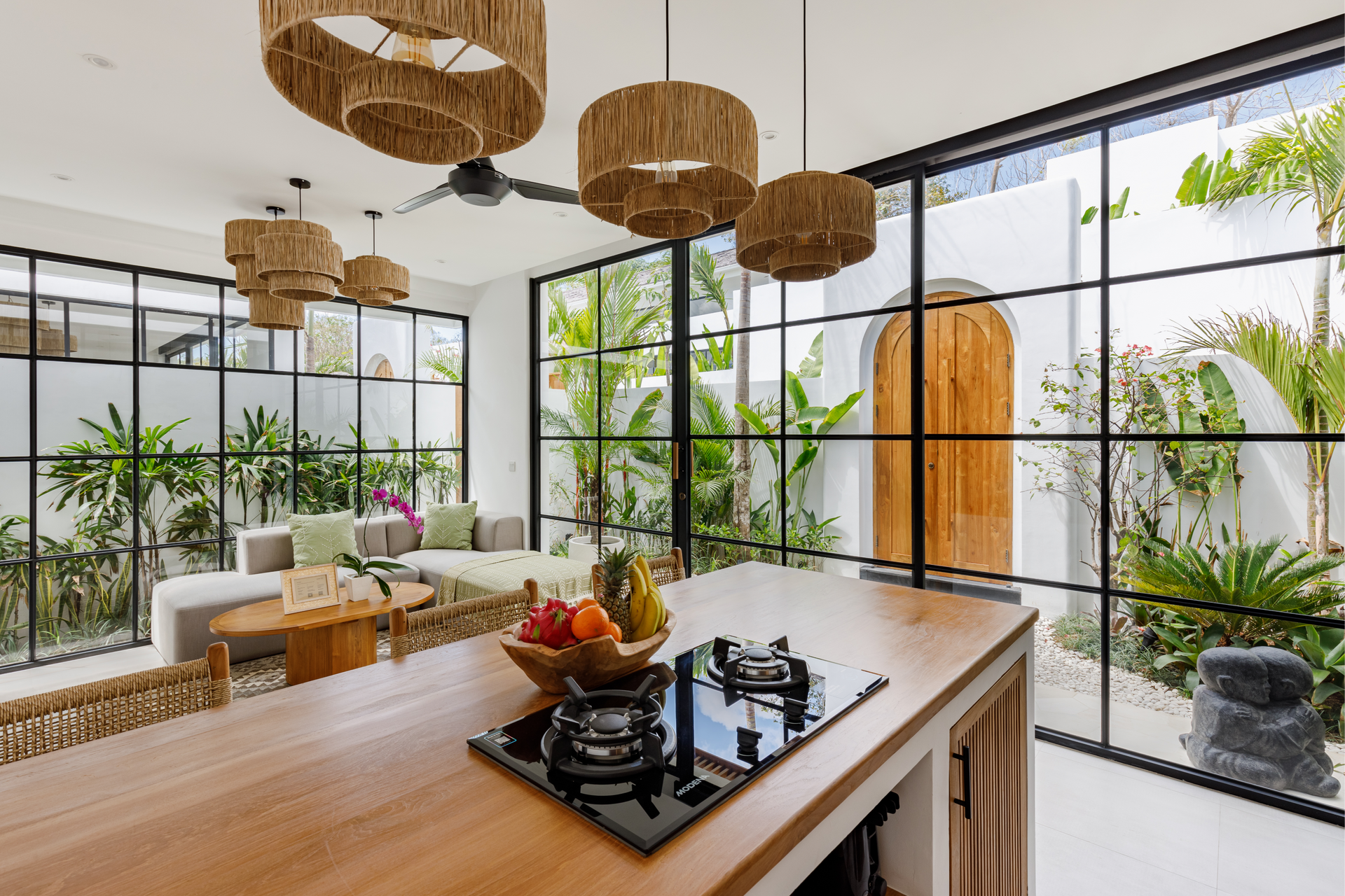 Bright modern kitchen with island, black cooktop, wicker pendant lights, and lush indoor plants by large windows