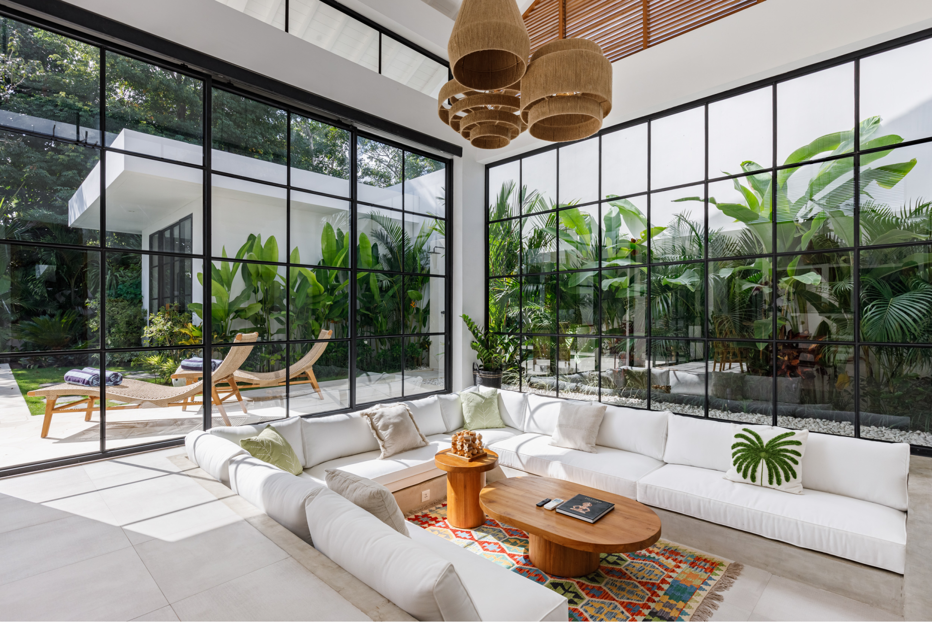 Bright modern living room with white sofas, wood coffee table, and floor-to-ceiling tropical garden windows