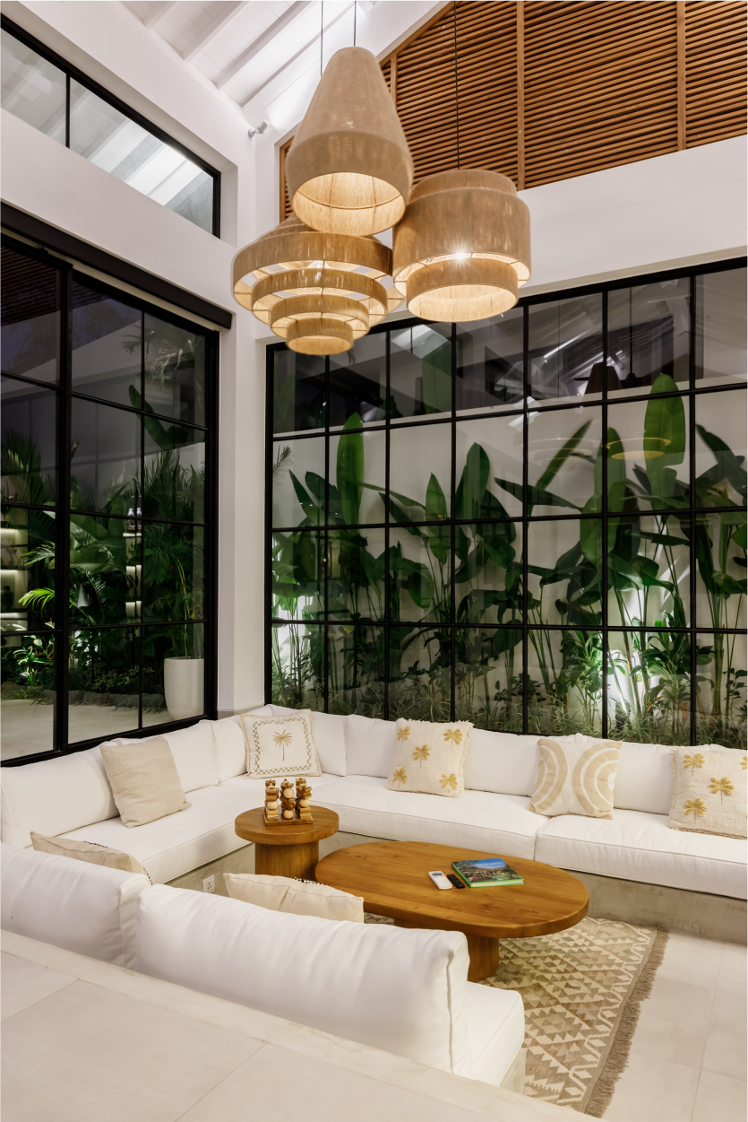 Bright modern lounge with white sectional seating, woven pendant lights, and tropical greenery behind glass walls
