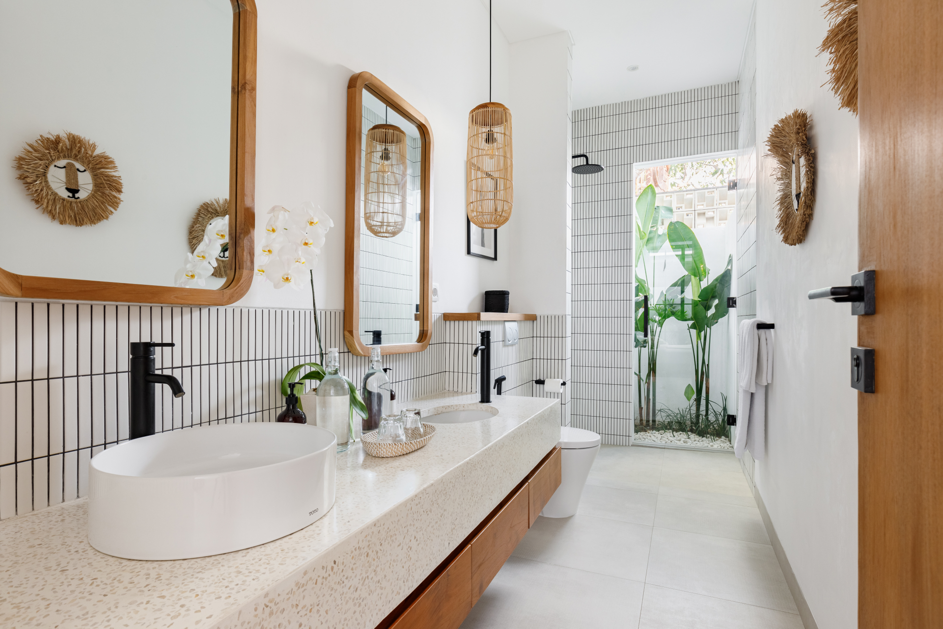 Bright modern bathroom with double sinks, mirror, wooden accents, and tiled walls, opening to a leafy view.