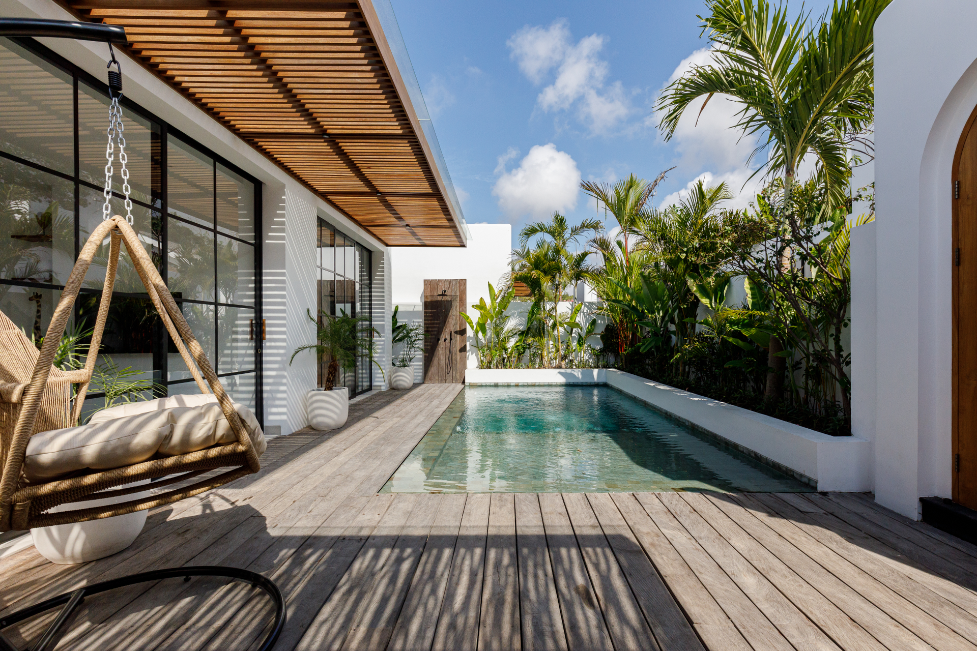 Modern villa patio with a hanging chair, wood deck, and narrow pool beside lush tropical greenery