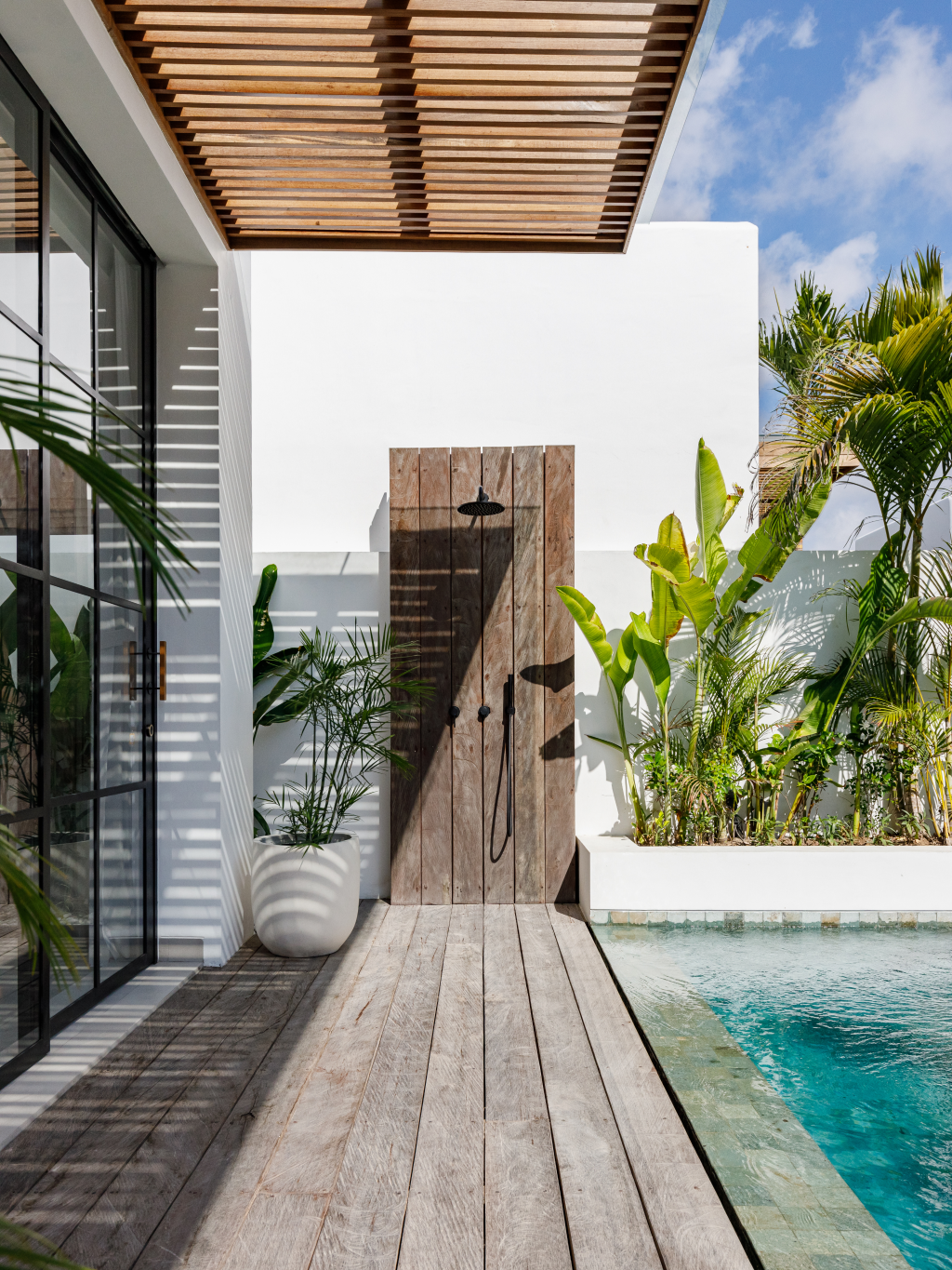 Modern patio with wood deck, white walls, tropical plants, and a turquoise pool under a slatted pergola