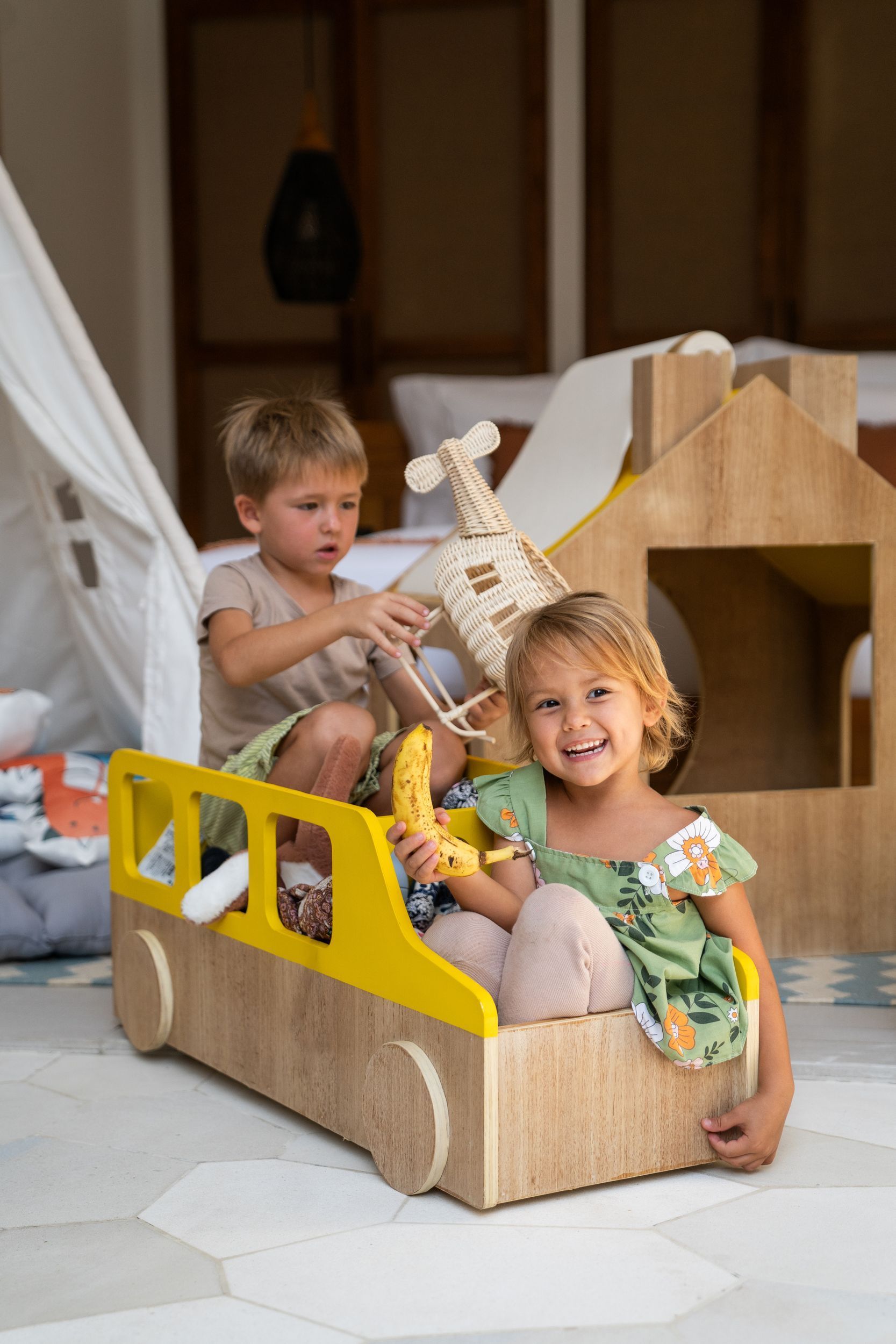 Two children playing in a cardboard toy car indoors, one smiling in front and one seated behind