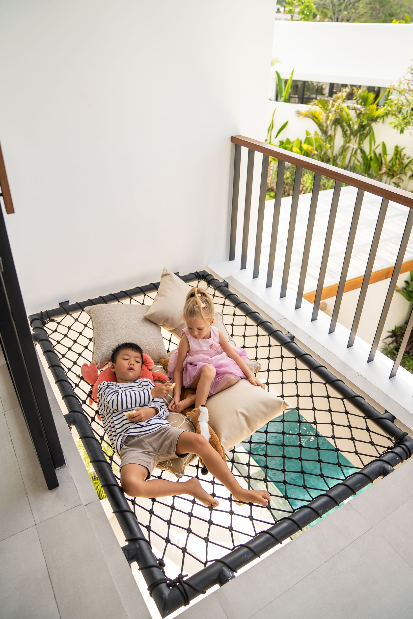 Two children lying on a balcony hammock, one in red and white stripes and one in pink, with plants nearby