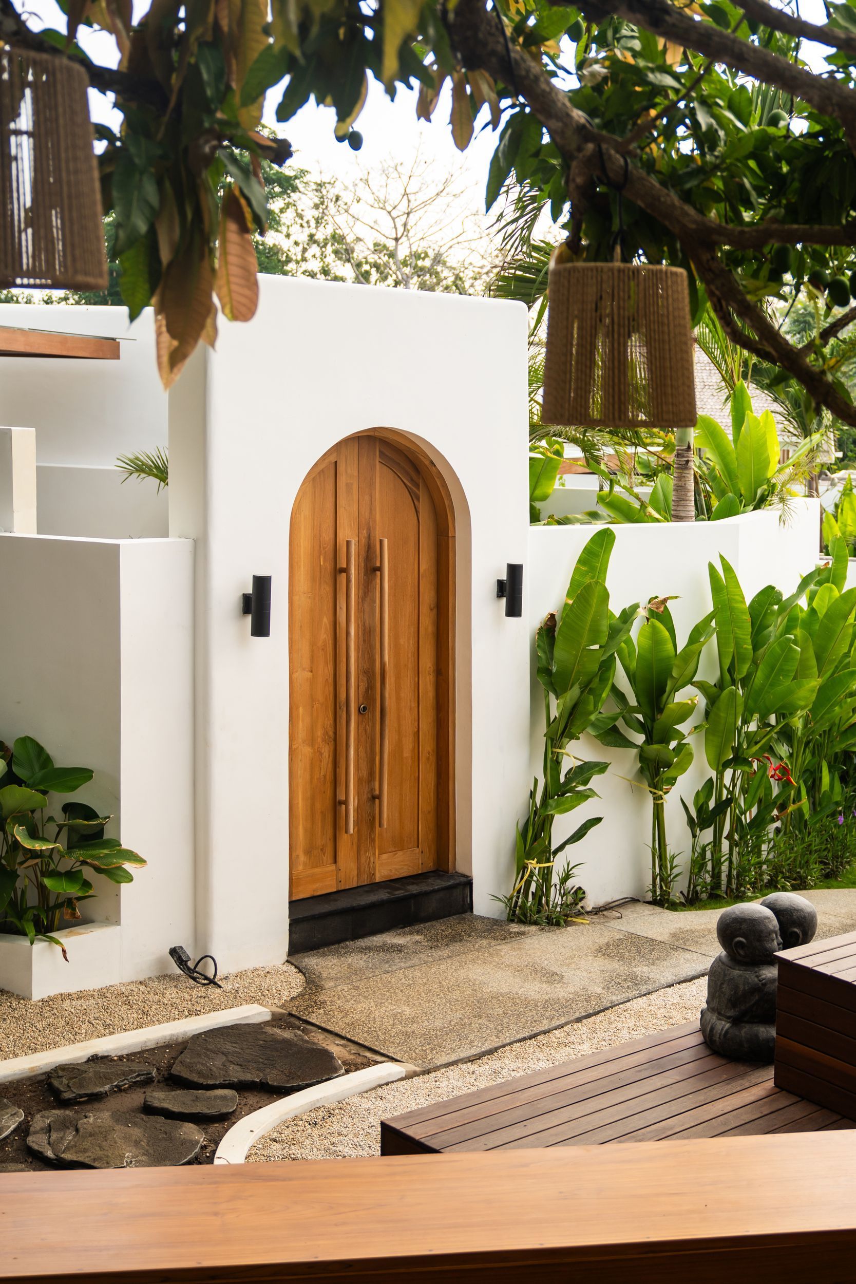 Tropical courtyard with white walls, wooden arched door, and lush greenery around a stone pathway