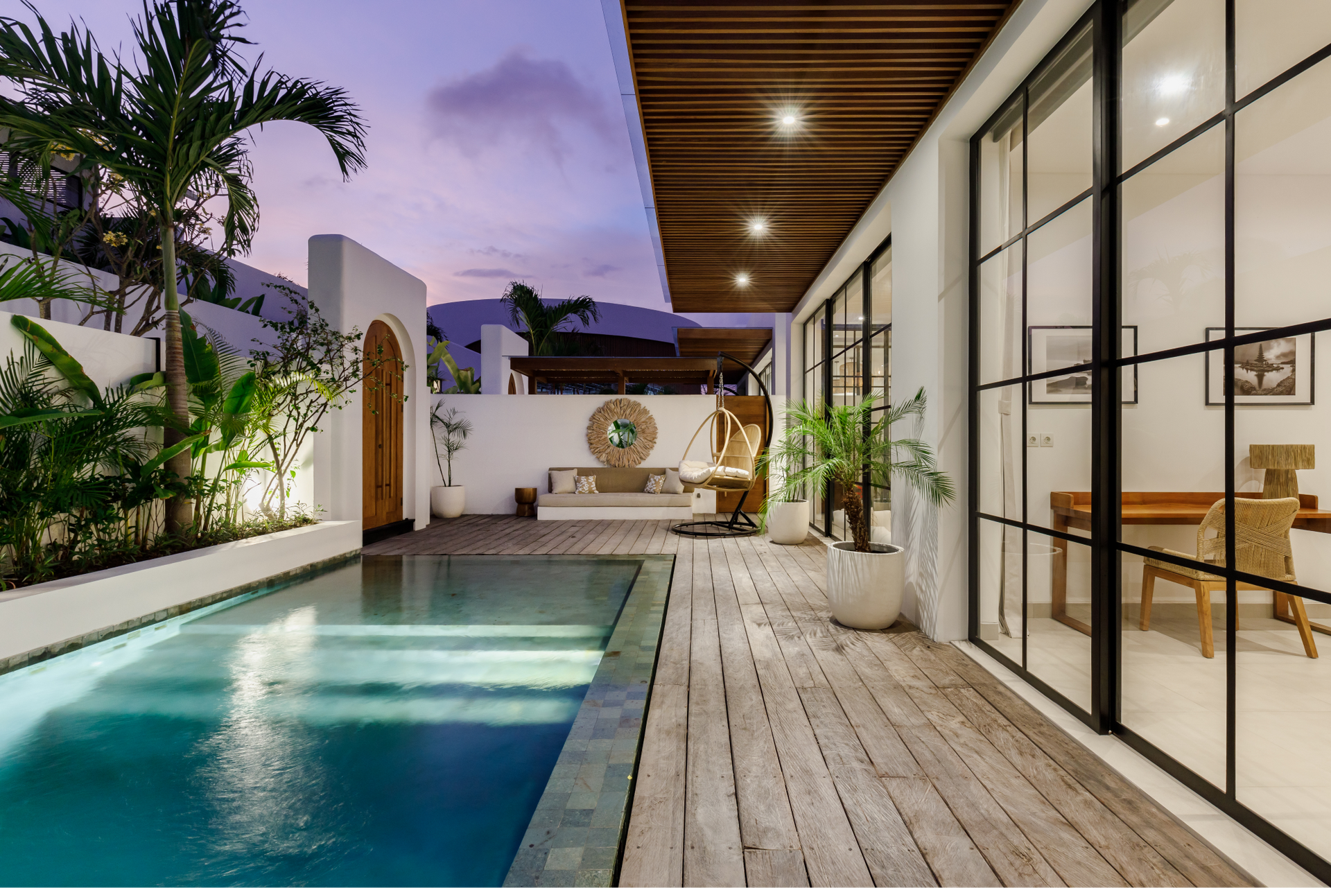Modern backyard patio with a narrow pool, wood deck, and glass-walled lounge at dusk