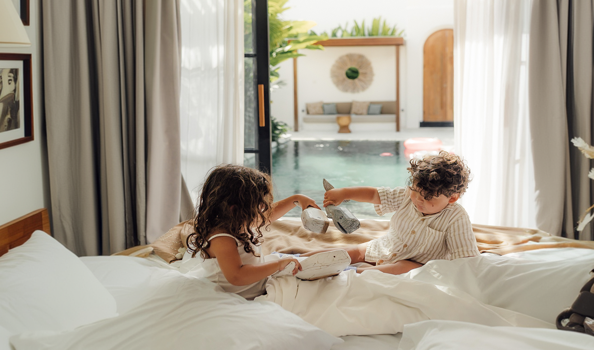 Two children play on a bed in a bright bedroom with open curtains and a sunny patio beyond.