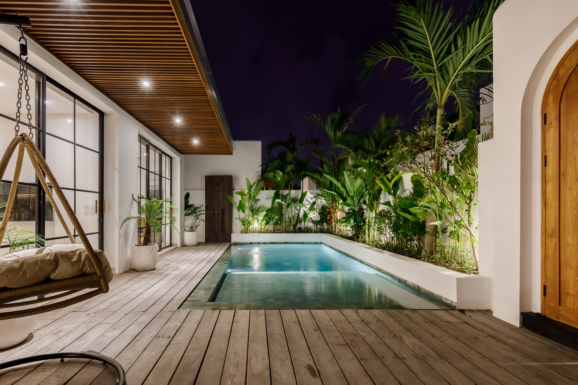 Modern backyard patio with a lit swimming pool, wooden deck, and tropical plants at night