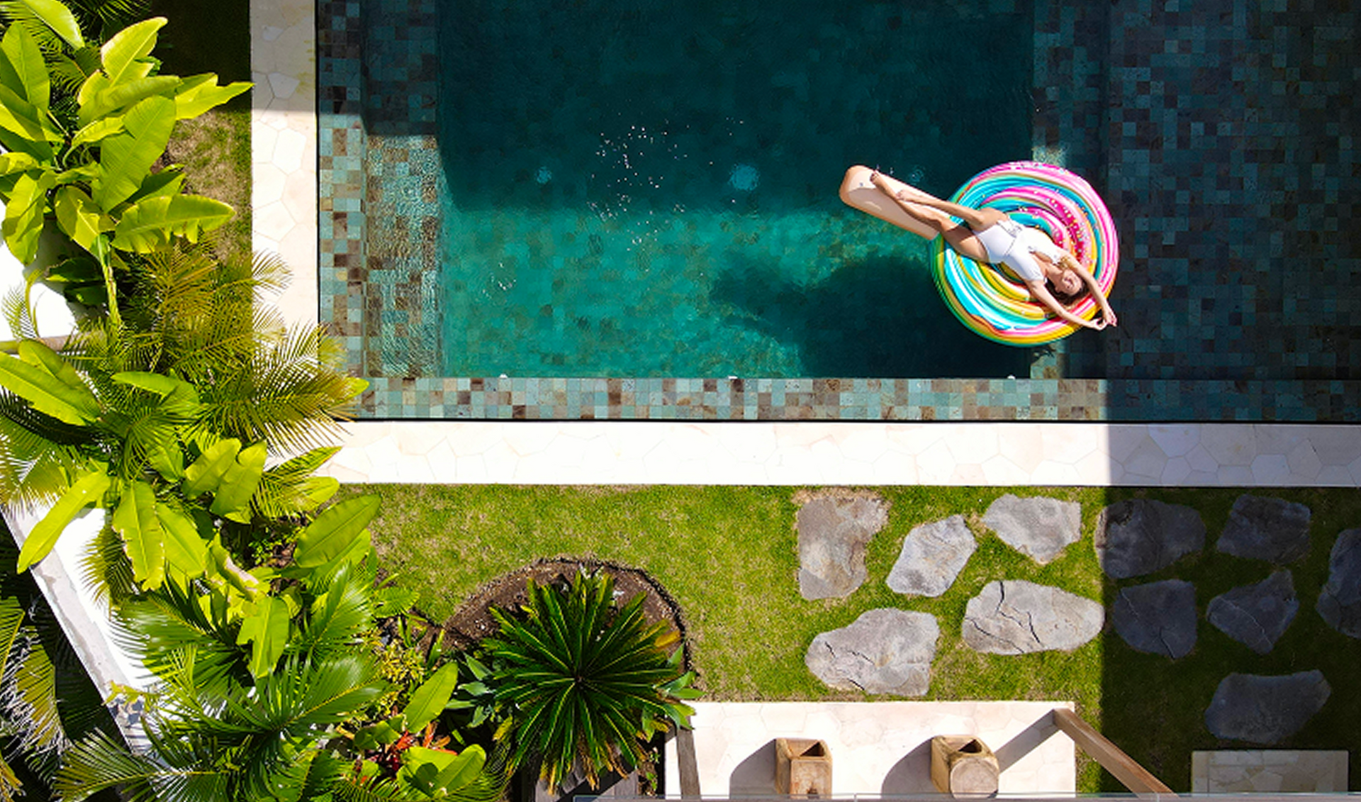 Top-down view of a person floating in a turquoise pool beside a stone path and lush greenery