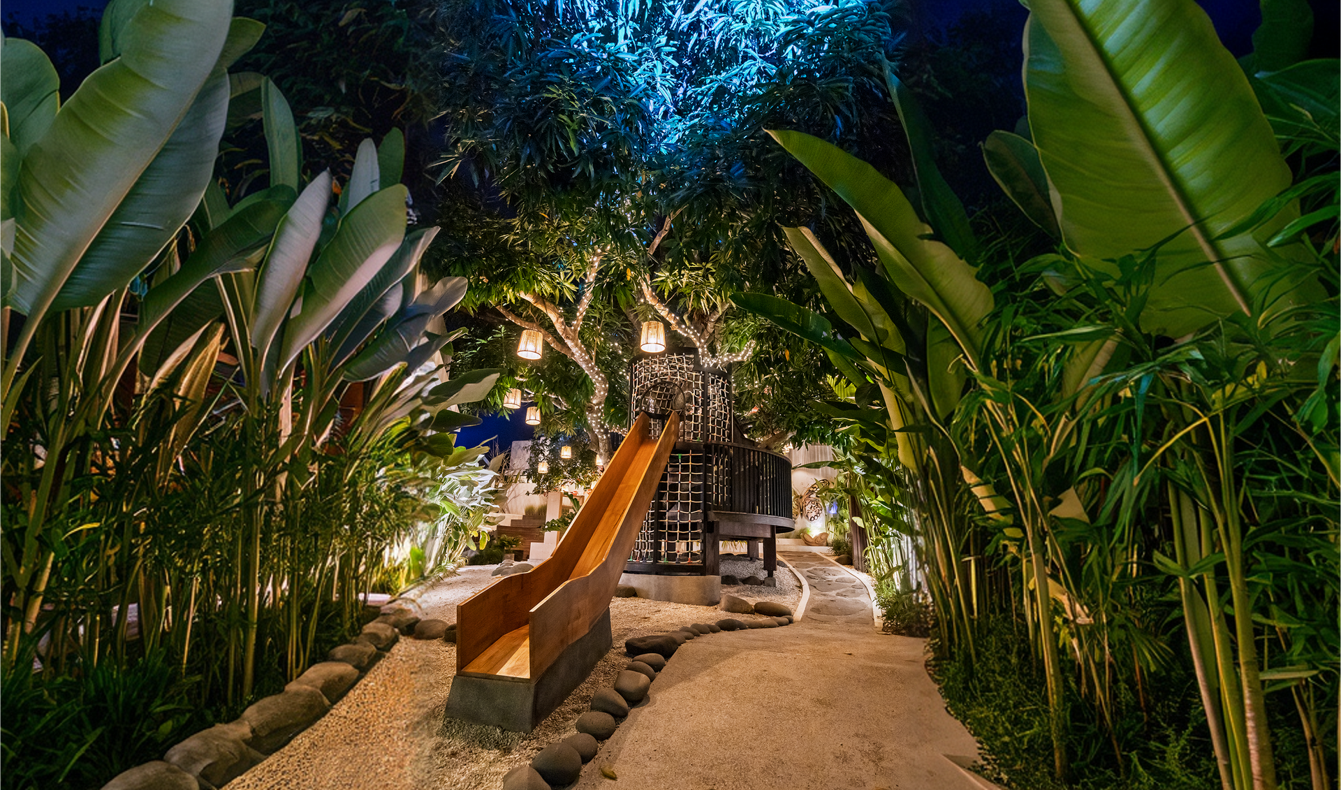 Garden pathway at dusk with lush green plants, warm lights, and a wooden bench beside a table.