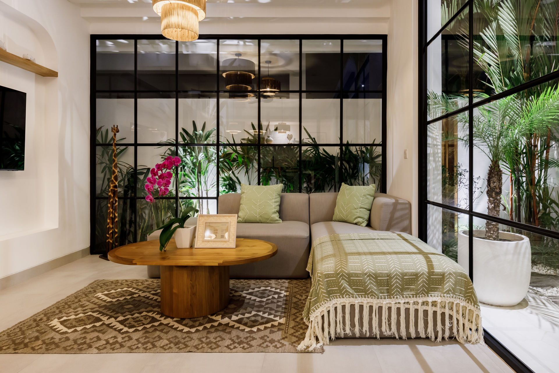 Modern living room with beige sectional, wooden coffee table, plants, and black-framed glass partition