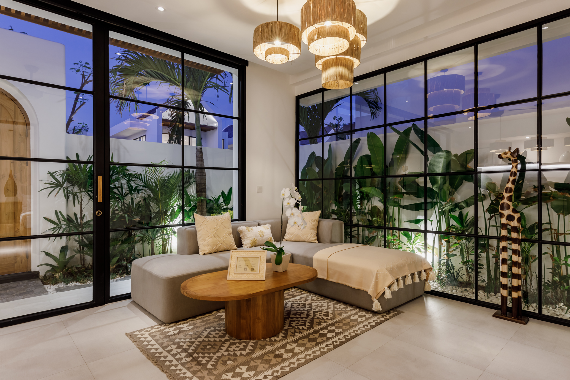 Modern living room with large glass walls, beige sectional, wooden coffee table, plants, and giraffe decor