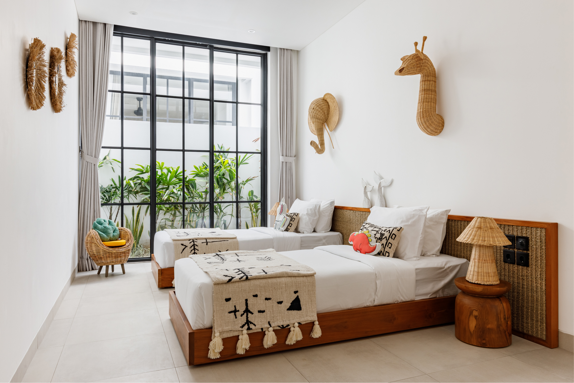 Bright boho bedroom with two beds, woven wall decor, and a large window overlooking greenery