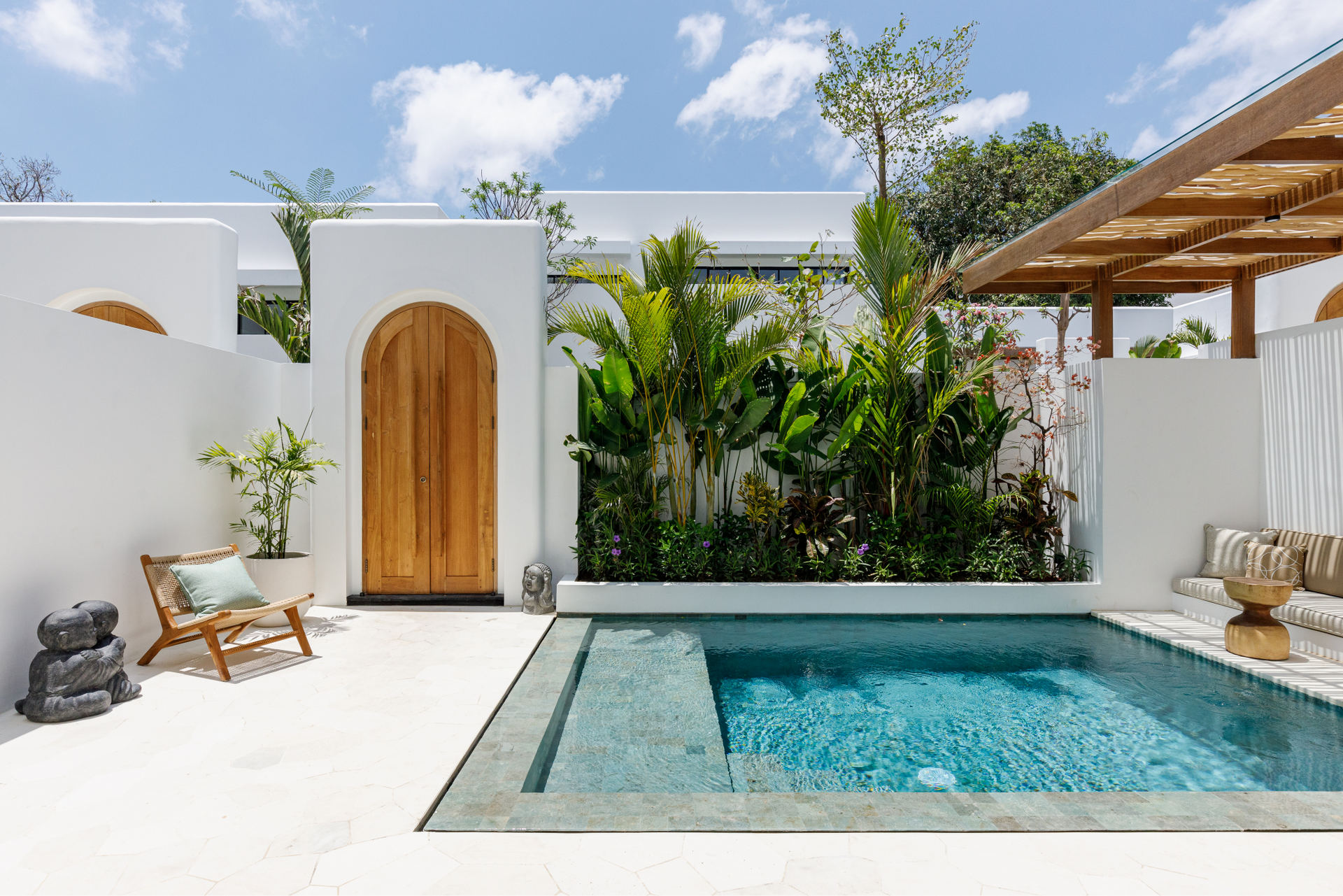Modern white patio with small turquoise pool, wooden door, lounge chairs, and lush tropical plants under a blue sky