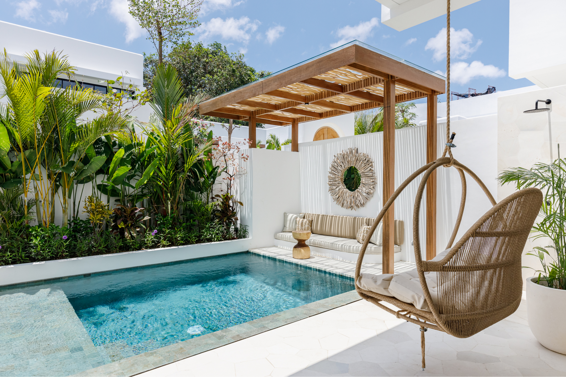 Bright patio with a small blue pool, hanging chair, and wooden pergola beside lush greenery