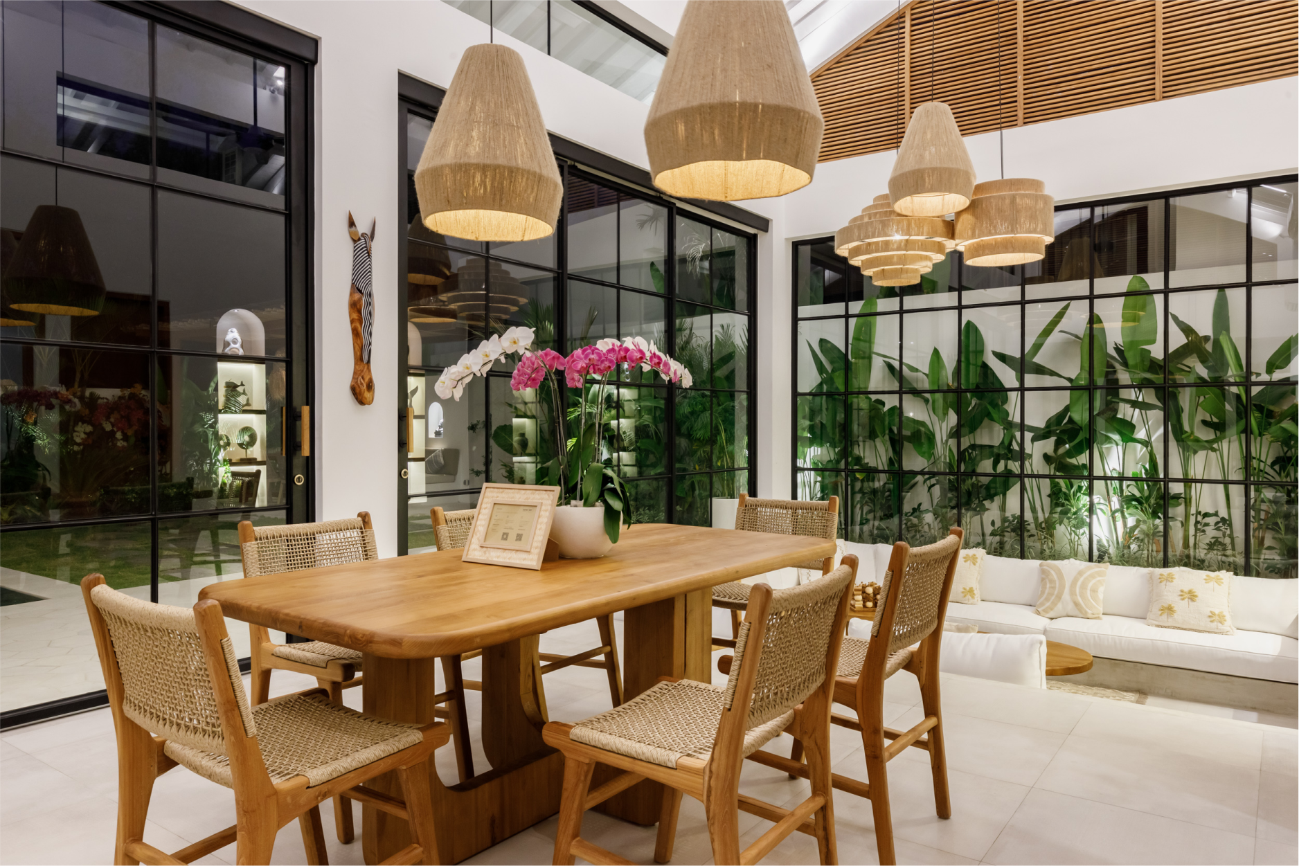 Bright modern dining area with a wooden table, wicker chairs, hanging lamps, and indoor plants