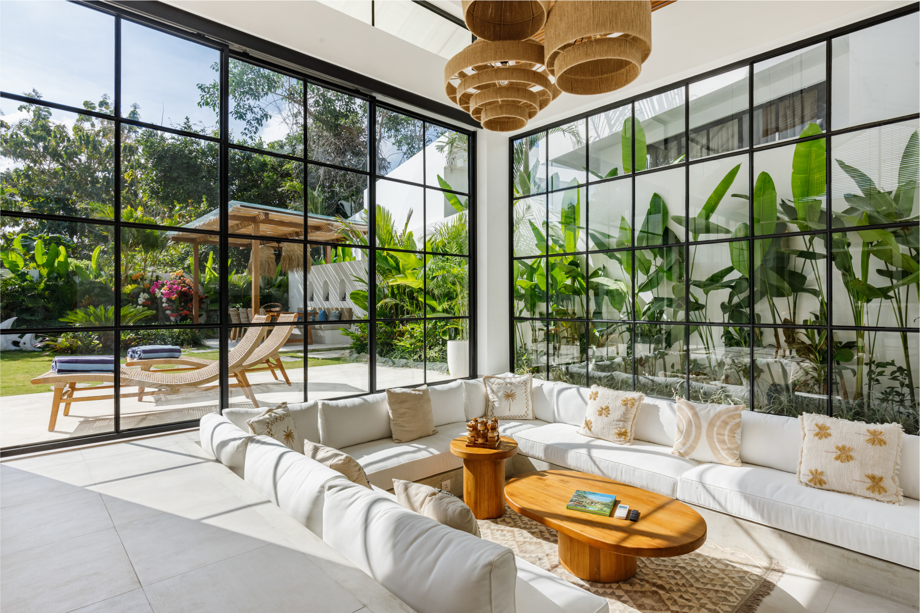 Bright sunroom with white sofa, wood coffee table, and large black-framed windows overlooking lush greenery.
