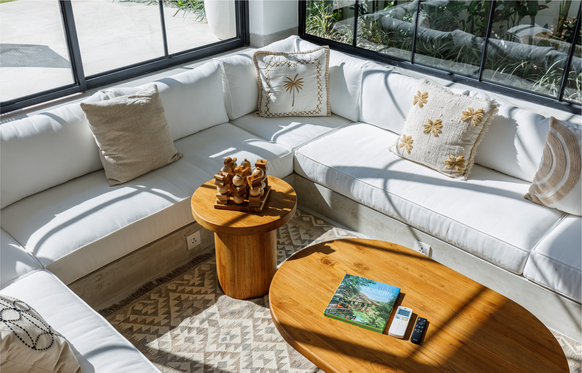 Sunlit white sectional sofa by windows with gold-accent pillows, wooden tables, and a patterned rug