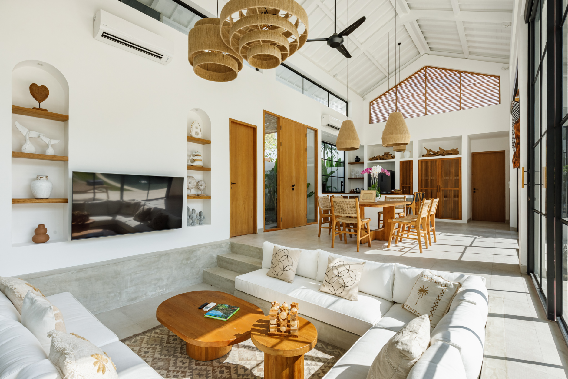 Bright modern living room with white sofas, wooden dining table, hanging lights, and large glass doors